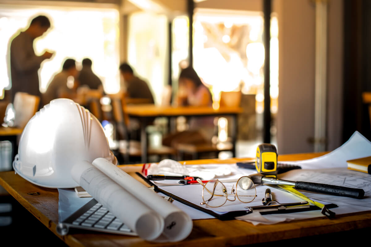 Foreground of a hard hat, blueprints, and other objects against a blurred background of people around a conference table